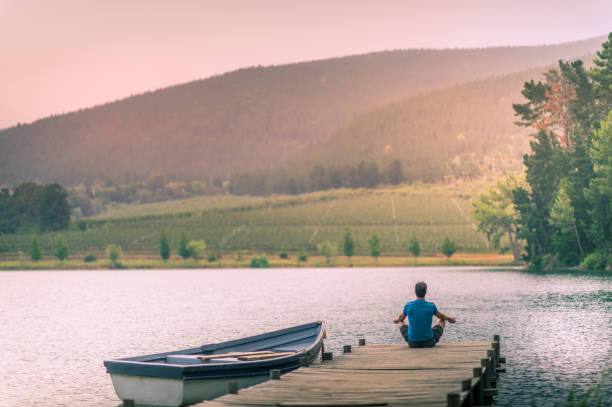 man meditating on a wooden jetty on a beautiful lake with a moored rowboat at sunset - loch rowboat lake landscape imagens e fotografias de stock