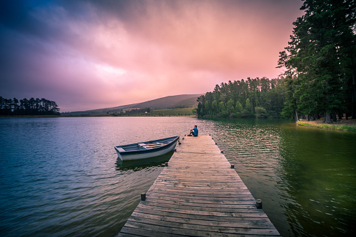 Wide shot of a 40-44-year-old Caucasian man on a wooden jetty on a full lake of water overlooking mountains and woodlands with a moored rowboat with a dramatic purple orange coloured mood Cape Winelands South Africa