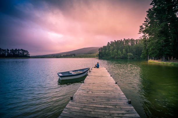 a mid adult man sitting on a wooden jetty in a beautiful lake with a moored rowboat dramatic sky - loch rowboat lake landscape imagens e fotografias de stock