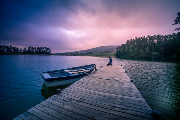 wide shot of a mid adult man on a wooden jetty kneeling praying in a beautiful lake with a moored rowboat dramatic sky - loch rowboat lake landscape imagens e fotografias de stock