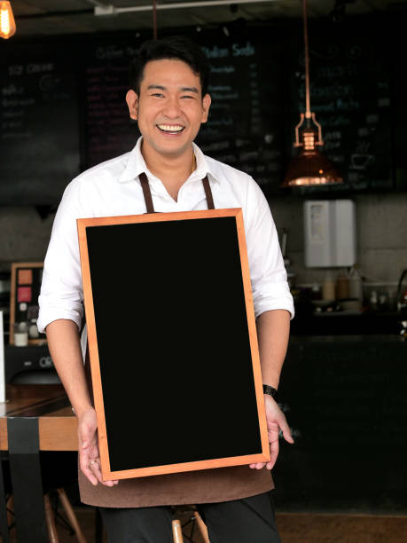コーヒーショップの前でブランクブラックボードを保持しているアジアのバリスタマン。 - black sign holding vertical ストックフォトと画像