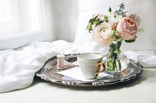 Spring, summer still life scene. Cup of coffee, blank greeting card, envelope on old silver tray at windowsill. Vintage feminine styled photo, floral composition, pink English roses and Ranunculus.