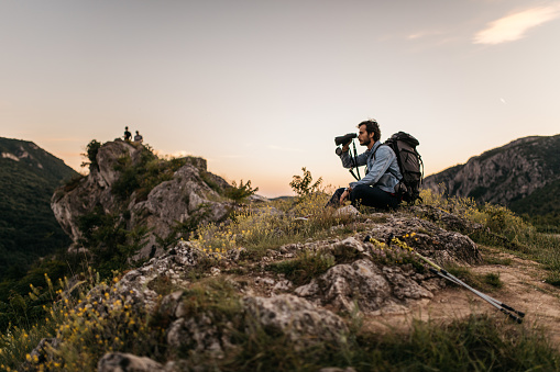 One man, hiker sitting high on mountain alone, holding binoculars, enjoying the view.