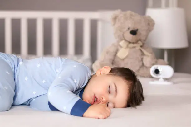 Photo of Cute little baby boy in light blue pajamas sleeping peacefully on bed at home with baby monitor camera and soft teddy bear toy at background. Child daytime sleeping schedule