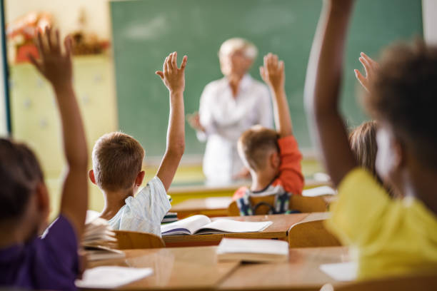 istock Rear view of elementary students raising their hands on a class at school. 1128876873