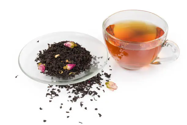 Prepared black tea in the glass cup and dried leaves of black tea with Chinese rose buds and petals of orange flowers in saucer and scattered beside to him on a white background