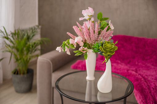 two white vases on round glass coffee table in stylish living room with grey scandinavian sofa. Modern bright interior.Vase of plant,flowers,succulent plant and palm tree in pot on the background. Home decoration interior.