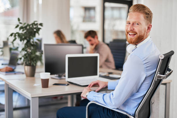 Smiling CEO sitting in office in formal wear and looking over shouder at camera. Smiling CEO sitting in office in formal wear and looking over shouder at camera. On desk laptop, coffee and smart phone. In background employees working. founder stock pictures, royalty-free photos & images