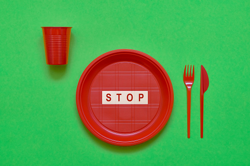 Disposable red plastic plate with word STOP inside, red knife and fork on green background. Top view, copy space. Plastic free concept. No Use symbol