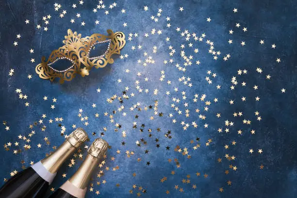 Two champagne bottles, golden carnival mask and confetti stars  on blue background. Flat lay of Christmas, anniversary, New Year, Purim, Carnival celebration concept. Copy space, top view.