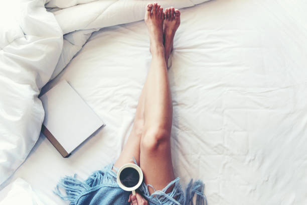 donne dalle gambe ravvicinate su letto bianco.  le donne che leggono libri e bevono caffè al mattino rilassano l'umore nella stagione invernale.   concetto di stile di vita. - bed human leg home interior comfortable foto e immagini stock