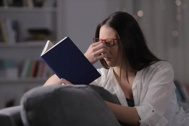 Lady with eyeglasses suffering eyestrain reading book in the night