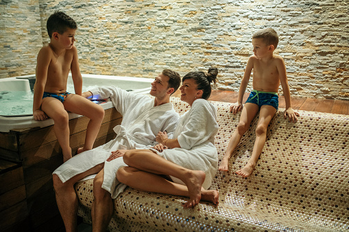Happy family with two children relaxing in spa while sitting on warm bench made of round mosaic tiles next to the hot bath jacuzi tube