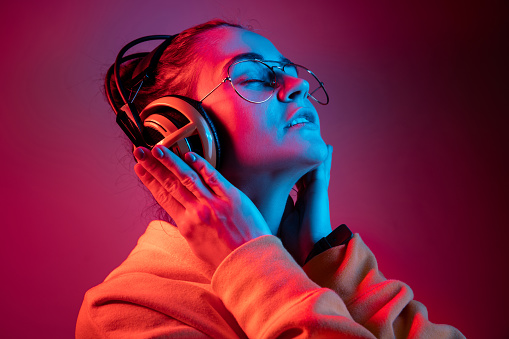 Fashion pretty woman with headphones listening to music over red neon background at studio.