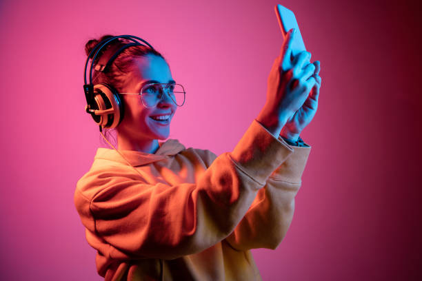 Fashion pretty woman with headphones listening to music over neon background Fashion pretty woman with headphones listening to music and making selfie photo over red neon background at studio. mobile phone lights up stock pictures, royalty-free photos & images