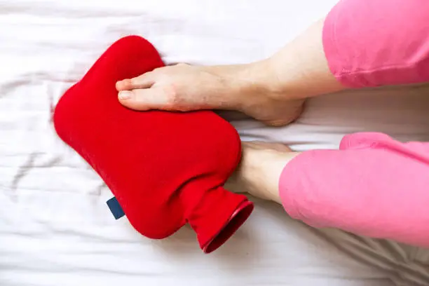 Top view of person with cold feet in bed on a red hot water bottle.; Warming of cold feet.