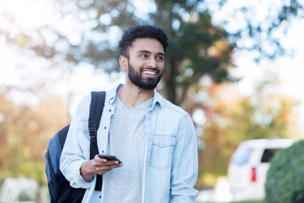 Confident male college student on campus Young adult male colleague student walks on campus. He is carrying a backpack and smartphone. indian subcontinent ethnicity stock pictures, royalty-free photos & images