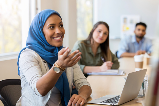 A cheerful Muslim businesswoman sits in front of a laptop at a conference table during a meeting.  She gestures as she shares ideas.