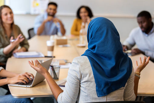 In this rear view, a Muslim businesswoman wearing a hijab sits at a conference table before a group of design coworkers.  She shrugs and gestures as she speaks.