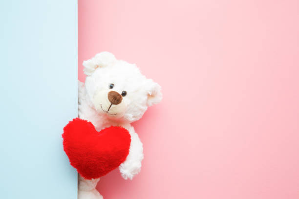 istock Smiling white teddy bear holding red soft heart and looking behind pastel blue wall. Mock up for happy, positive idea. Empty place for emotional, sentimental text, quote or sayings on pink background. 1128934083