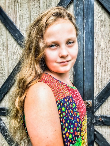 Beautiful Caucasian preteen girl, wearing a homemade patchwork quilt dress, and standing near a small shed or barn door, looking at the camera with confidence and taking a break outdoors on her hobby farm. Portrait mode with selective focus.