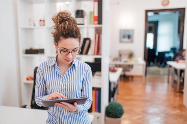 istock Elegant business woman standing at workplace, using digital tablet, front view. 1128944723