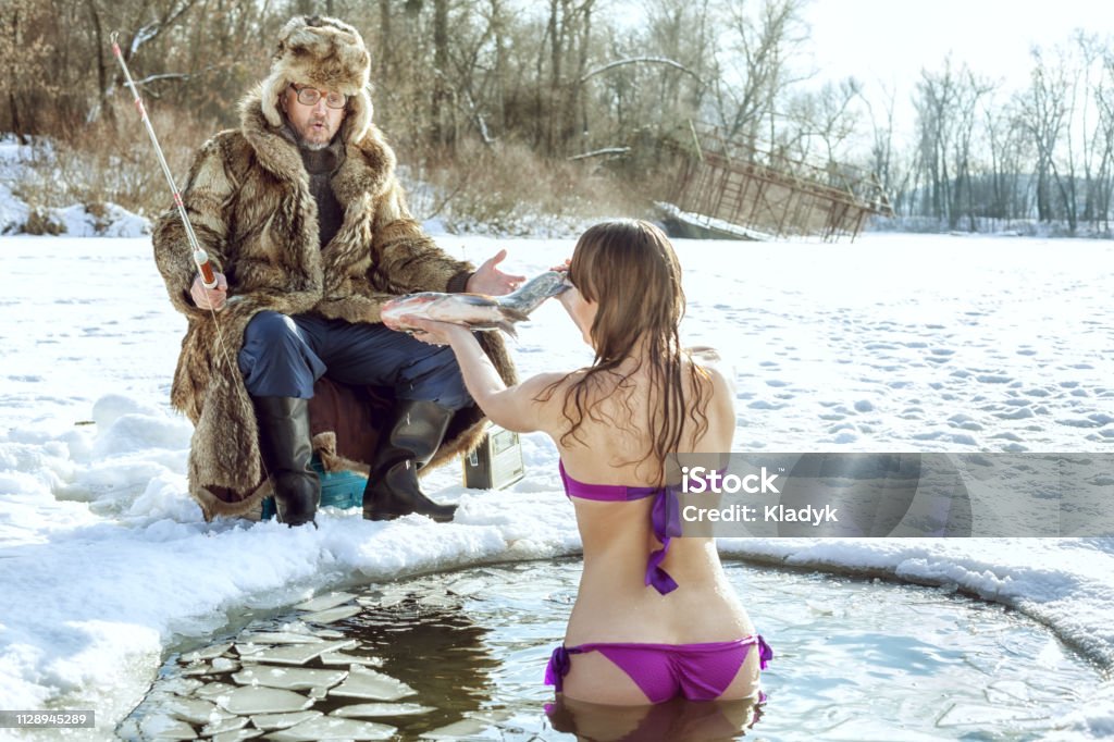 Elderly man is sitting at the hole in the ice and fishes. Elderly man is sitting at the hole in the ice. Girl in swimsuit stands in ice-hole and hands him a fish. Fishing Stock Photo