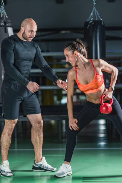 istock Instructor Assisting Young Woman Lifting Kettlebell At Gym 1128948234
