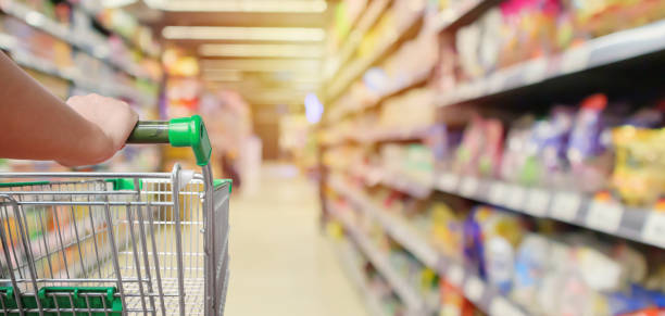 shopping cart in supermarket aisle with product shelves interior defocused blur background shopping cart in supermarket aisle with product shelves interior defocused blur background groceries stock pictures, royalty-free photos & images