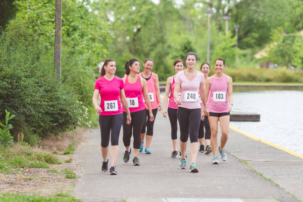 A group of female runners wearing pink prepare to go on a cancer fundraising run together Group of women runners in pink shirts and racing bibs gather in solidarity and get ready to run in a fundraising event. They are walking cheerfully toward the starting line. walking for a cause stock pictures, royalty-free photos & images