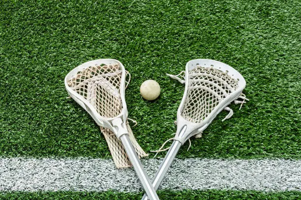 Looking down on a pair of Lacrosse sticks and a white ball sitting on the white Midfield-line of an artificial turf sports field.