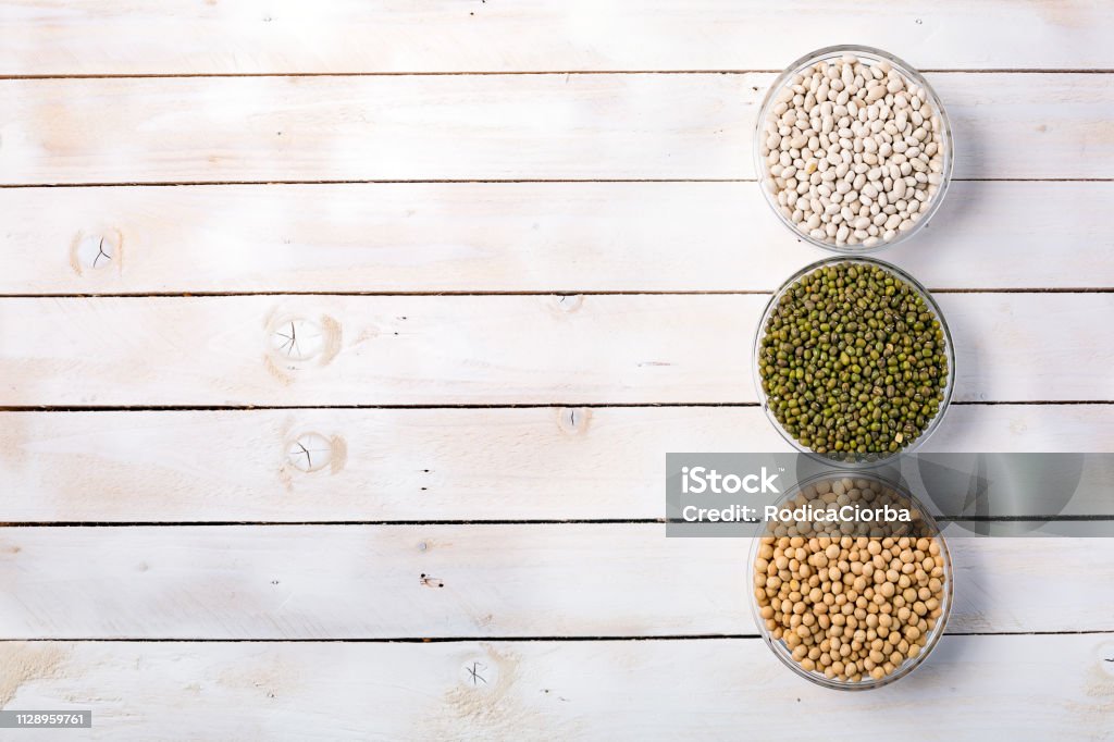 Raw dry various vegetable protein on white wood background. Raw mung beans, white beans and soybeans in glass bowls on white wooden background top view, space for text. Vegan protein source. Superfoods and Healthy food clean eating. Above Stock Photo