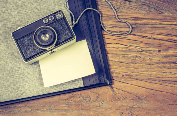 Travel memories concept: Flat lay of old camera and photo album with white blank paper for message. Vintage image with old photo camera, empty paper and black photo album on rustic wooden background. old black leather photo album stock pictures, royalty-free photos & images
