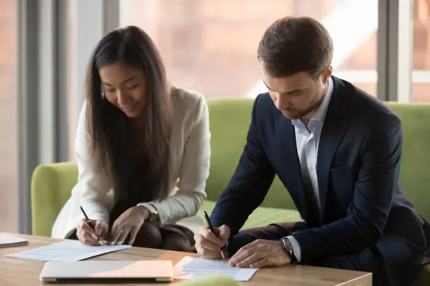 Asian businesswoman and caucasian businessman sign contracts documents at meeting, diverse male female business partners client and service provider put signature on legal papers making agreement