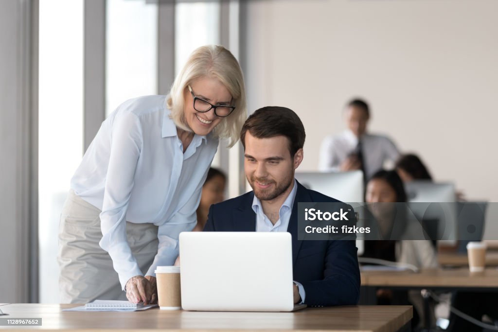Friendly mentor training employee in office helping with computer work Friendly middle aged female mentor executive training employee in office helping with computer work, smiling old corporate manager supervise motivating teaching new worker colleague looking at laptop Teaching Stock Photo