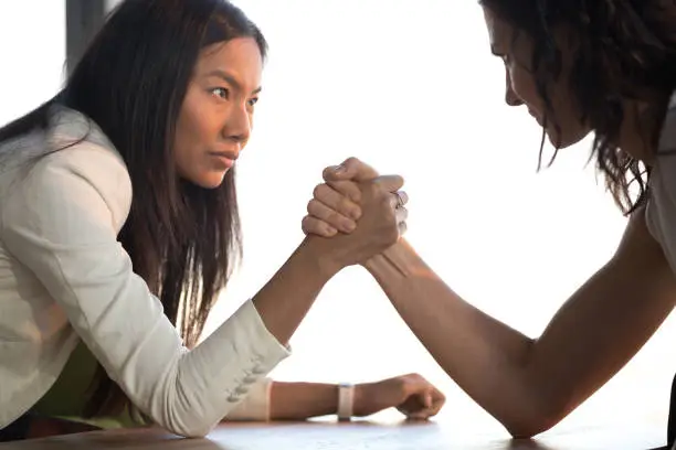 Two confident diverse businesswomen compete arm wrestling look in eyes feel jealous envious about success, young asian and caucasian female rivals armwrestling struggle for leadership rivalry concept