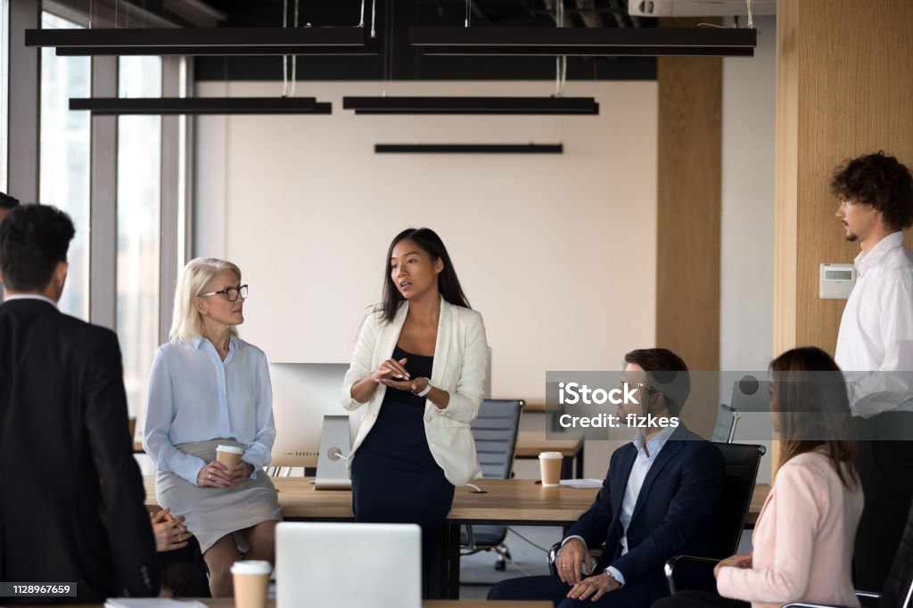 Serious asian coach speaking at diverse corporate group meeting Serious young female asian coach mentor team leader speaking at diverse corporate group meeting talking to office workers at lecture training teaching explaining presenting new business plan Leadership Stock Photo