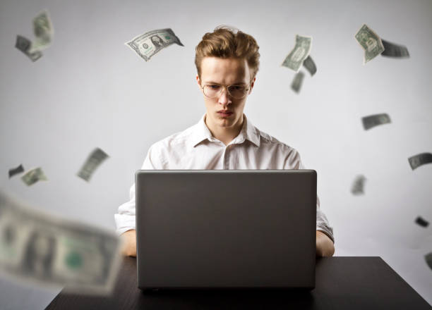 istock Young man using a laptop to browse the net. Young worker and falling dollar banknotes. Currency and job concept. 1128987381