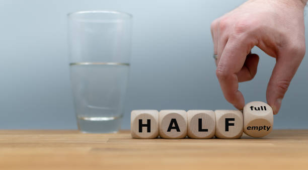 Hand turns a dice and changes the expression "half empty" to "half full". A half full glass of water is standing in front of a grey background. stock photo