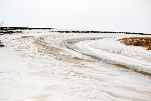 Snow on an S curve of a rural gravel road between a pair of tilled barren fields gathers enough soil to turn the snow drifts a shade of brown.  The accumulation leaves the road passable but difficult to travel