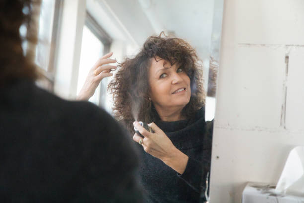 mature woman applying hair spray in front of mirror - woman in mirror backview imagens e fotografias de stock