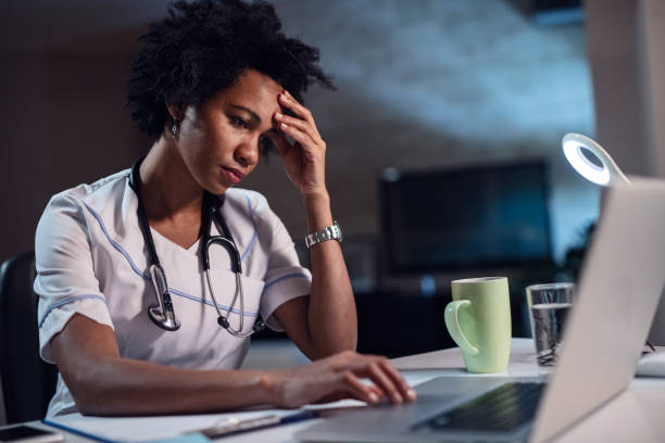 African American female doctor with headache working on laptop. Displeased black healthcare worker using computer and reading an e-mail at doctor's office. working overtime stock pictures, royalty-free photos & images