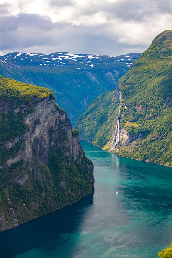 Mountain range and Seven Sisters Waterfall in the Geirangerfjord are captured on a cloudy summer day. The rocky mountains are covered with green trees and bushes. The landscape is mainly blue.