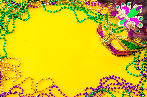 Fat Tuesday and Mardi Gras carnival concept theme with close up on a face mask full of color, feathers and texture and golden, green and purple beads isolated on yellow background with copy space