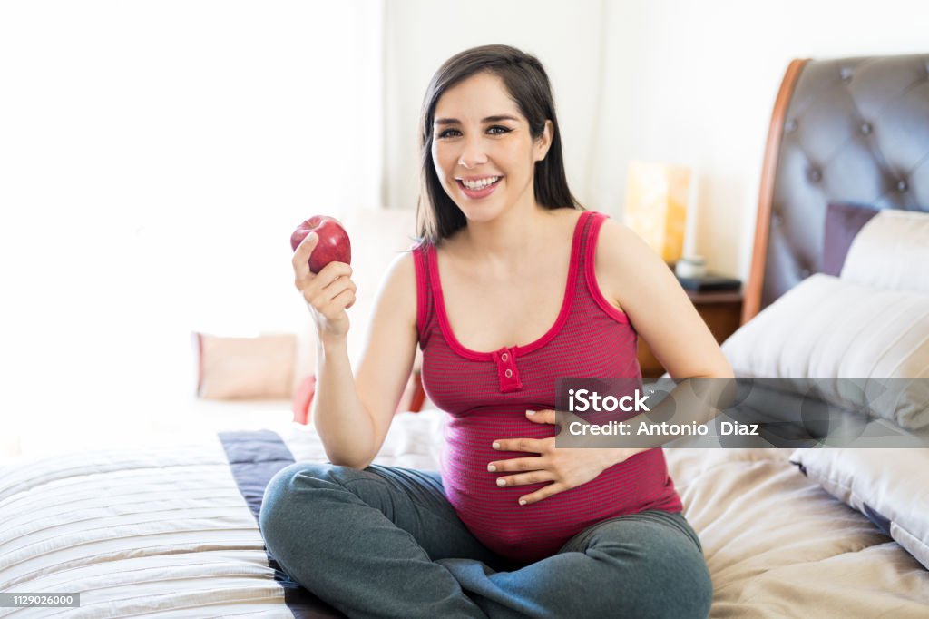 Expectant Woman Enjoy Snacking On An Apple Mother to be smiling while holding fresh apple while touching abdomen at home Eating Stock Photo