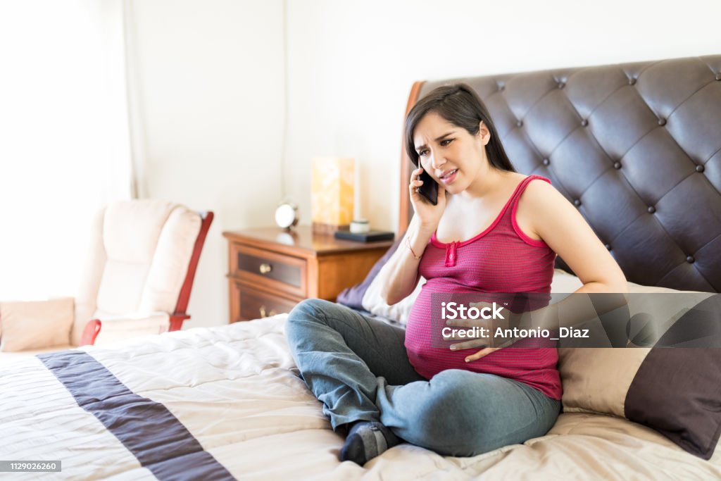 Pregnant Woman Feeling Contractions Calling For Help Mid adult woman in labor pain calling ambulance while sitting on bed Pregnant Stock Photo