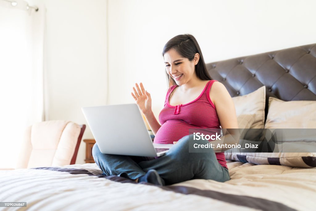 Sharing Pregnancy News Through Video Call Cheerful mother to be using conference calling application on laptop while relaxing on bed Pregnant Stock Photo