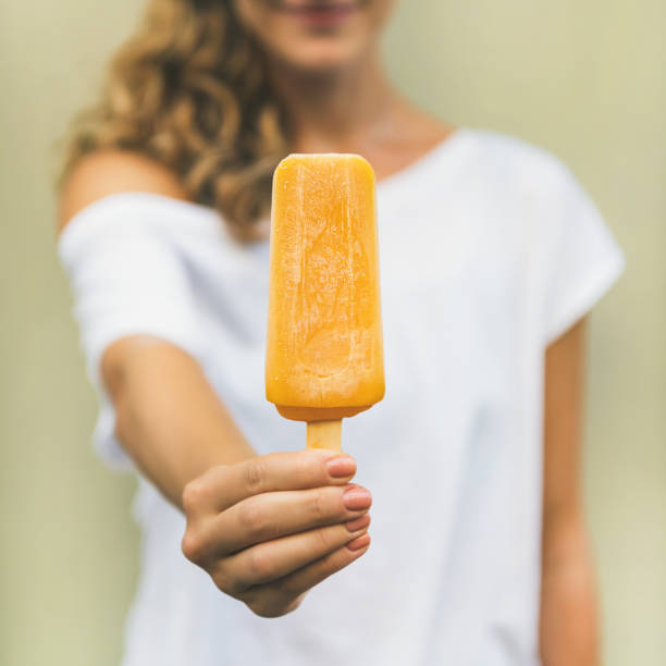 Healthy vegan orange mango citrus ice cream popsicle in hand of young woman with yellow wall at background, summer dessert and cheerful summer mood concept