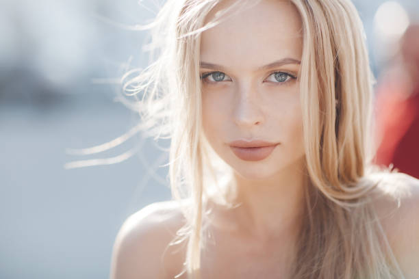 A young beautiful woman with long, straight hair and gray eyes, beautiful makeup, pink plump lips, sweet smile, blonde, spending her time outdoors in the summer in the city, dressed in a white jersey and checkered shirt, posing in the sunlight