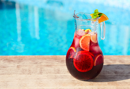 Glass jug of iced sangria with strawberry, orange, apple and lemon by the pool